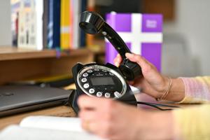 Schreibtisch mit Telefon und einer Hand, die den Telefonhöhrer hält - im Hintergrund in der Unschärfe eine Basisbibel und weitere Bücher.